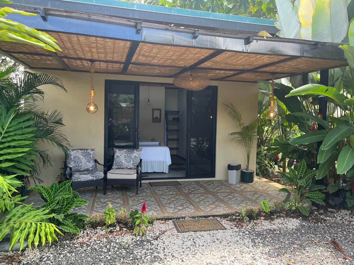 La entrada del bungalow de Reborn Spa bajo la pérgola de caña, con dos sillones y la camilla visible por la puerta corrediza
