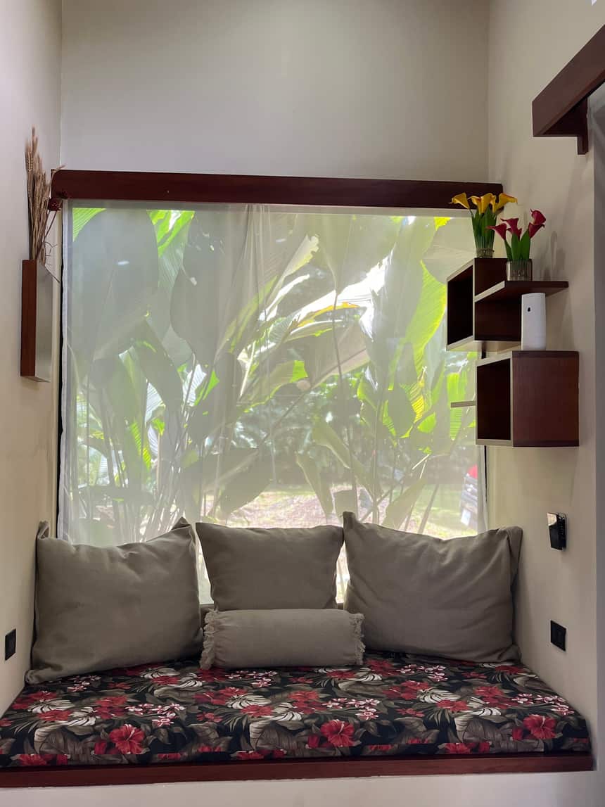 El diván bajo la ventana de Reborn Spa con cojines y almohadón floral, la vegetación al otro lado del vidrio