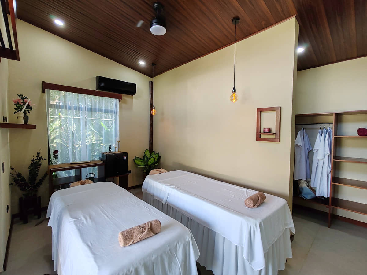 La sala de dos camillas de Reborn Spa: techo de madera, ventana al jardín, batas colgadas y estantería de madera