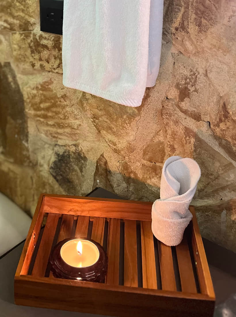 Bandeja de madera con vela encendida y toalla enrollada bajo el toallero del muro de piedra de Reborn Spa